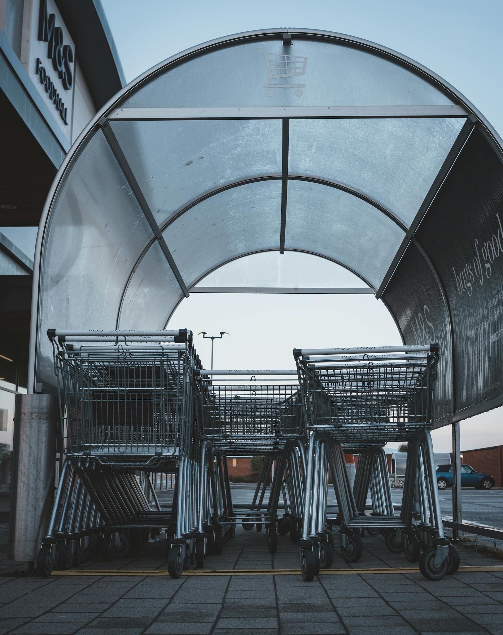 A group of metal shopping carts under a transparent shelter at a supermarket parking lot.